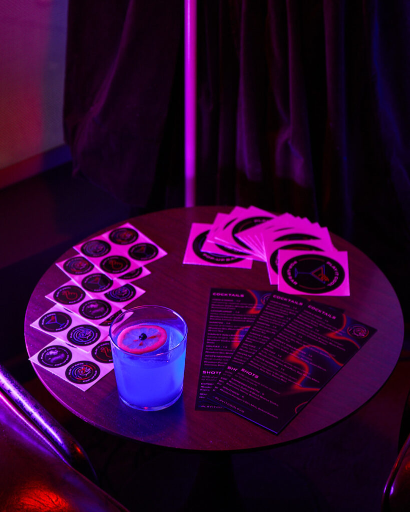 A blue cocktail with a lemon slice sits on a table with drink menus and stickers in a dimly lit setting.