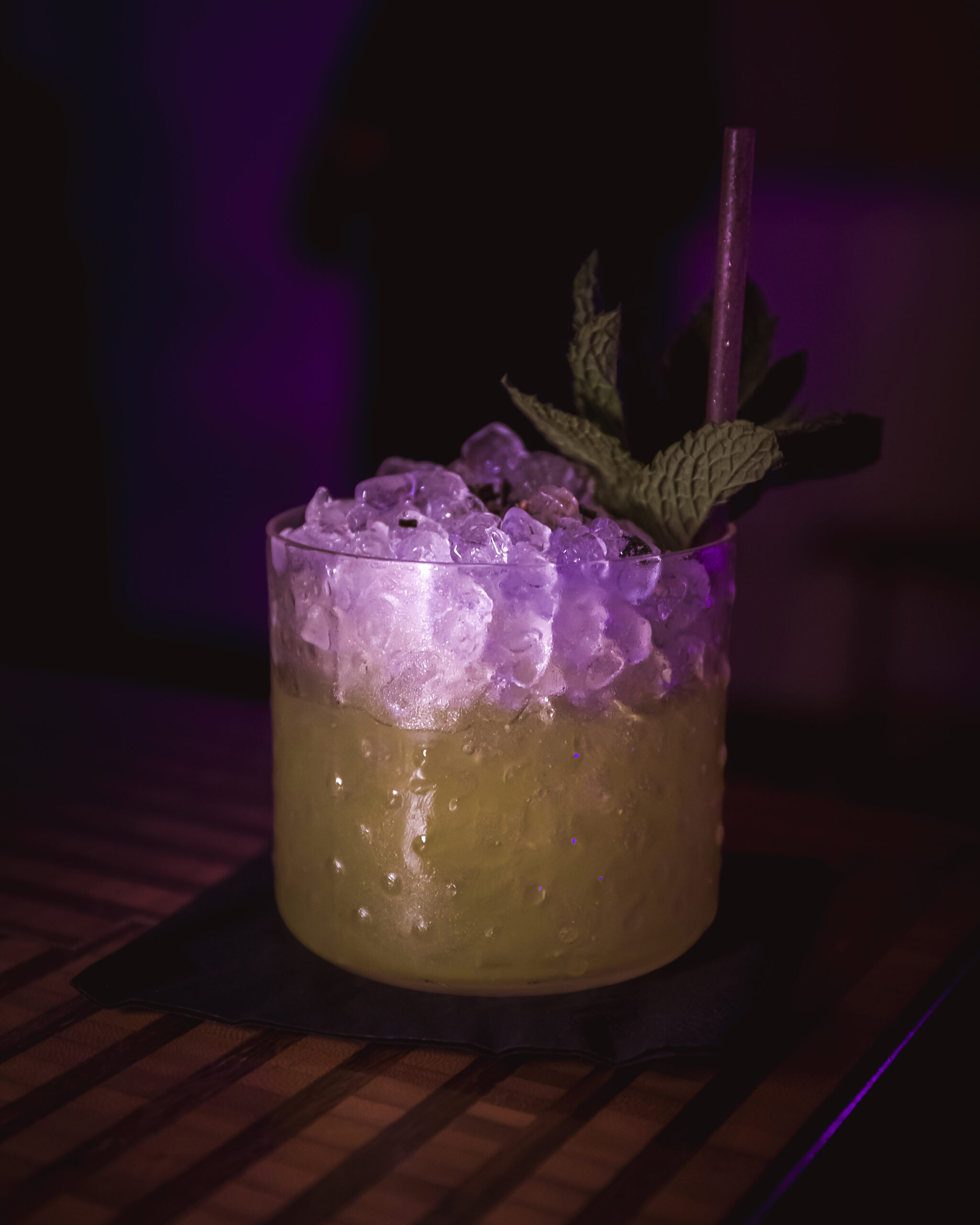 A Midori Sour cocktail served in a textured glass with crushed ice, garnished with mint leaves, under purple lighting.