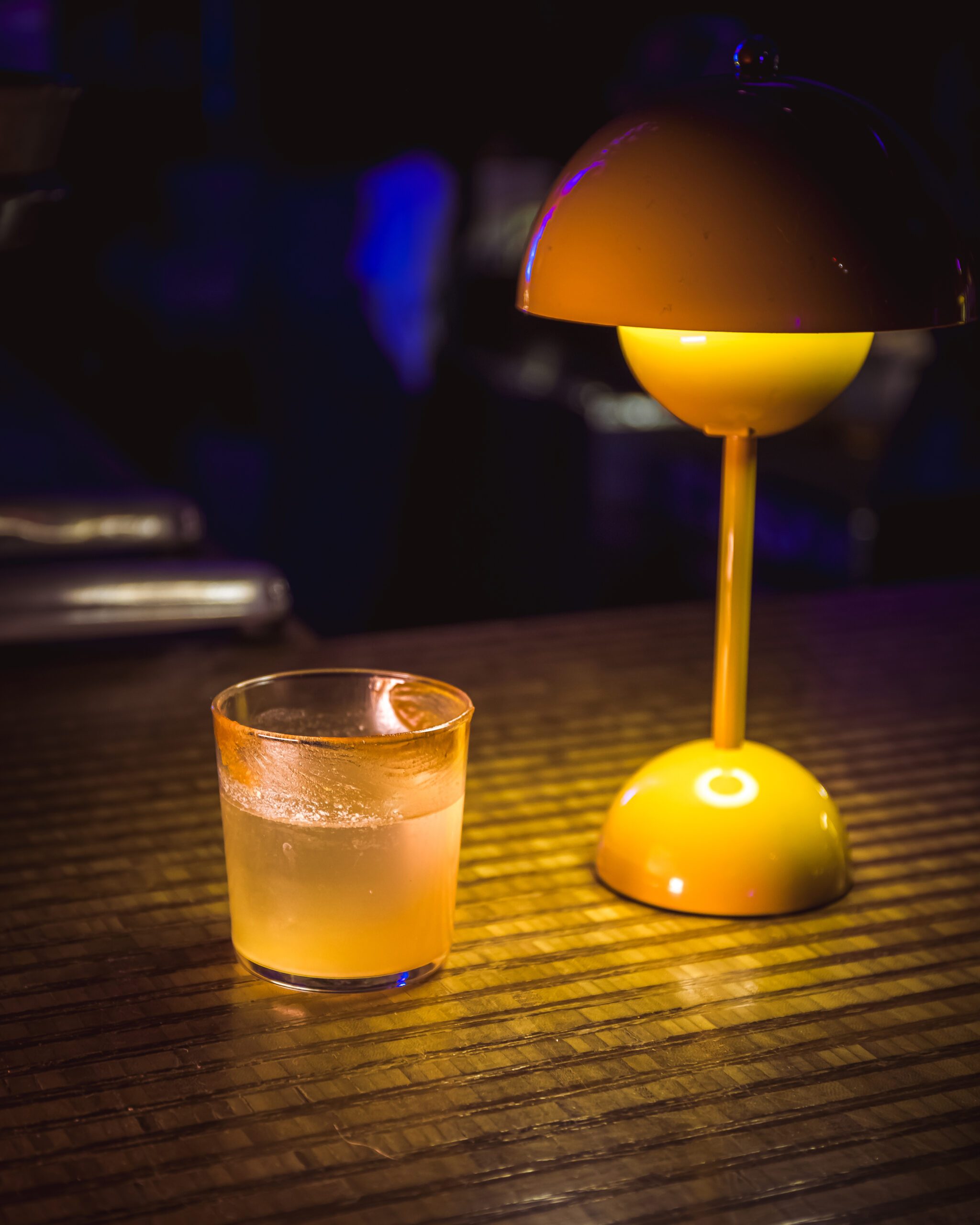 A glass of "Old Fashioned" cocktail sits on a wooden table next to a glowing yellow lamp, creating a warm ambiance.