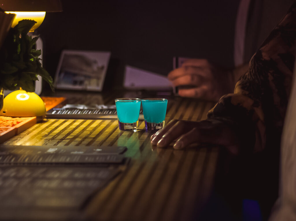 Two blue shots on a wooden bar counter at the Play It Cool event, with a hand and lamp visible in the background.