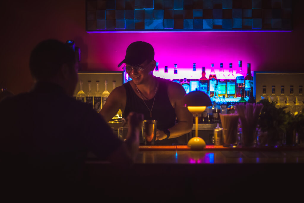Play It Cool Founder Parker Luthman smiles while bartending, illuminated by colorful lights.