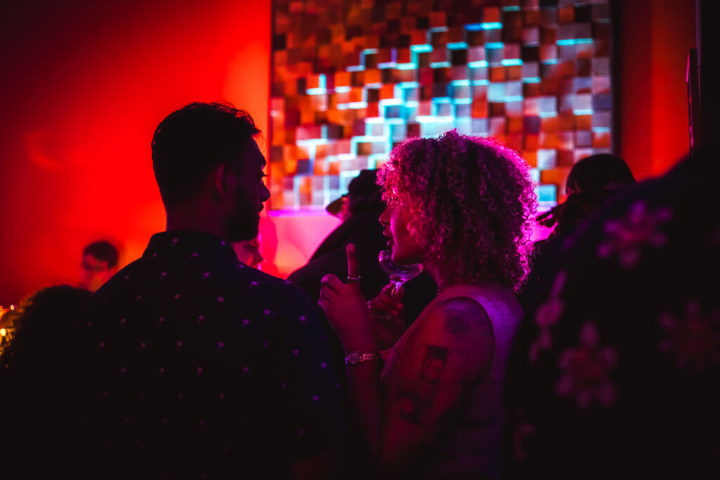 Two people converse in vibrant red lighting at the "Play It Cool Pop-Up Bar Event," featuring a colorful geometric backdrop.