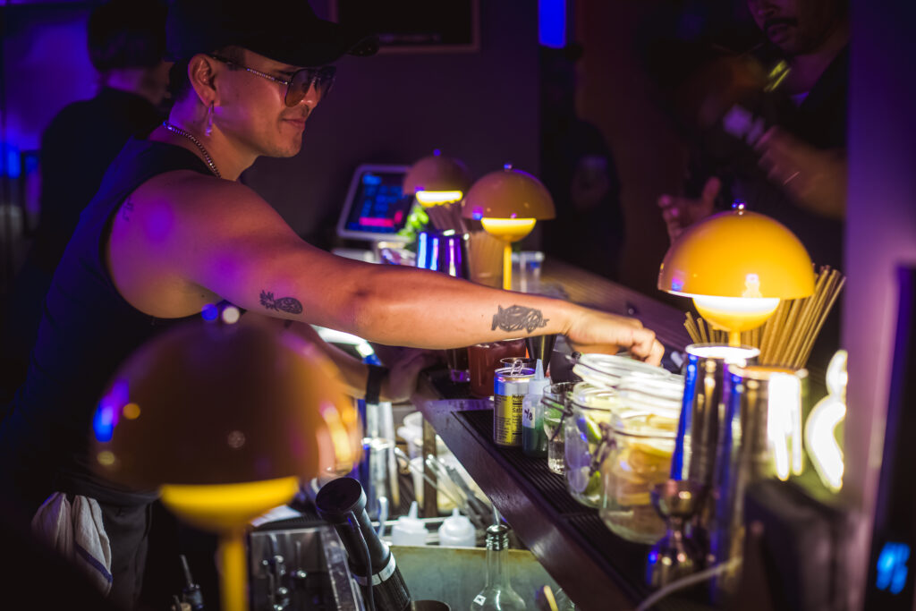 Play It Cool Founder Parker Luthman working behind the bar, surrounded by drinks and bar tools under warm lighting.