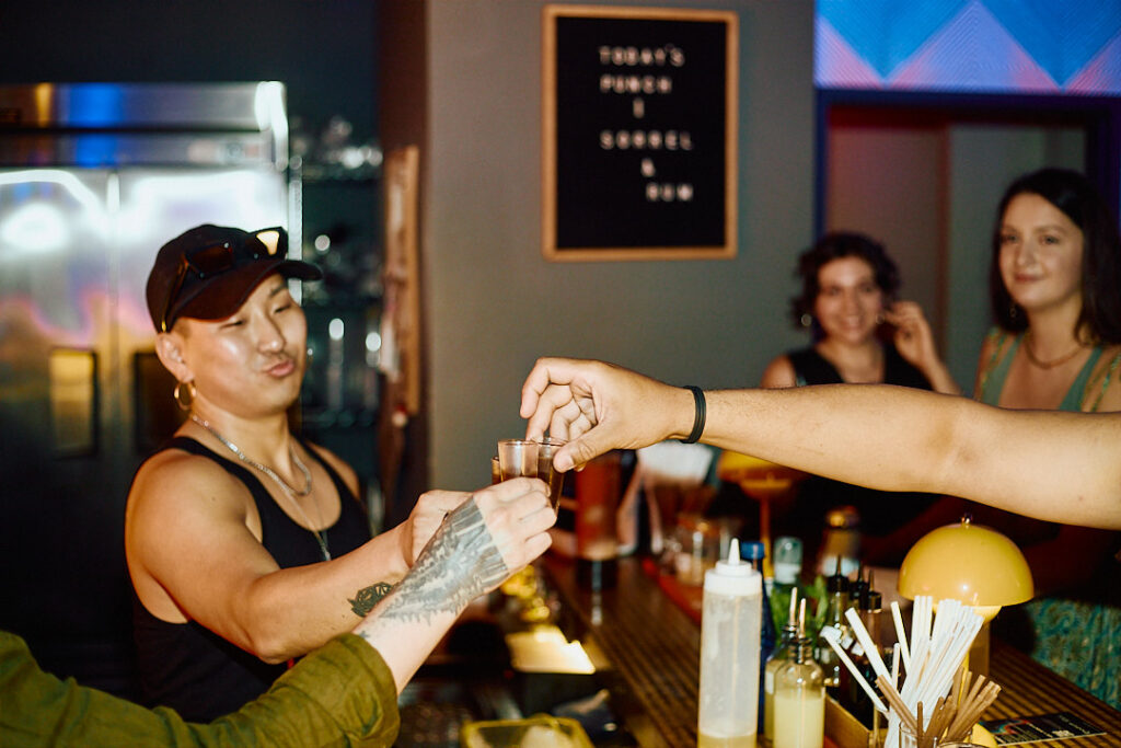A bartender hands a shot glass to a customer, while friends watch and smile in the background.