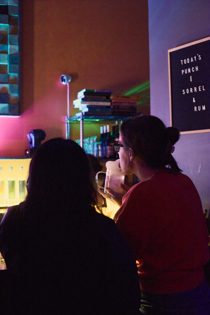Two people stand at a bar, one holding a drink, while colorful lights illuminate the scene.