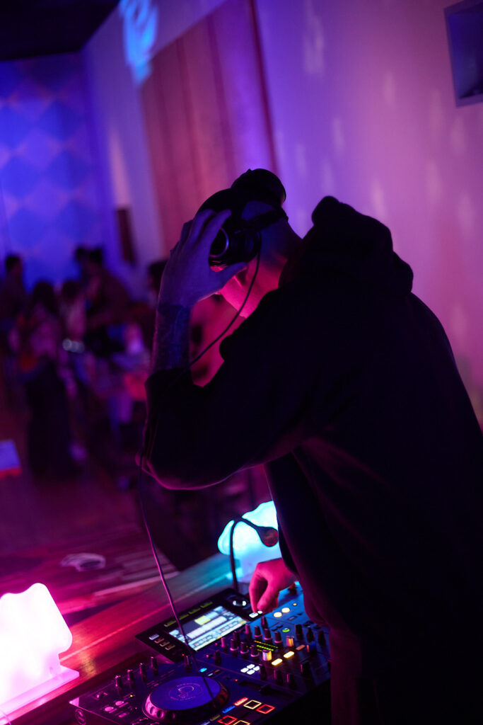 A DJ adjusts equipment under colorful lights, with people dancing in the background.