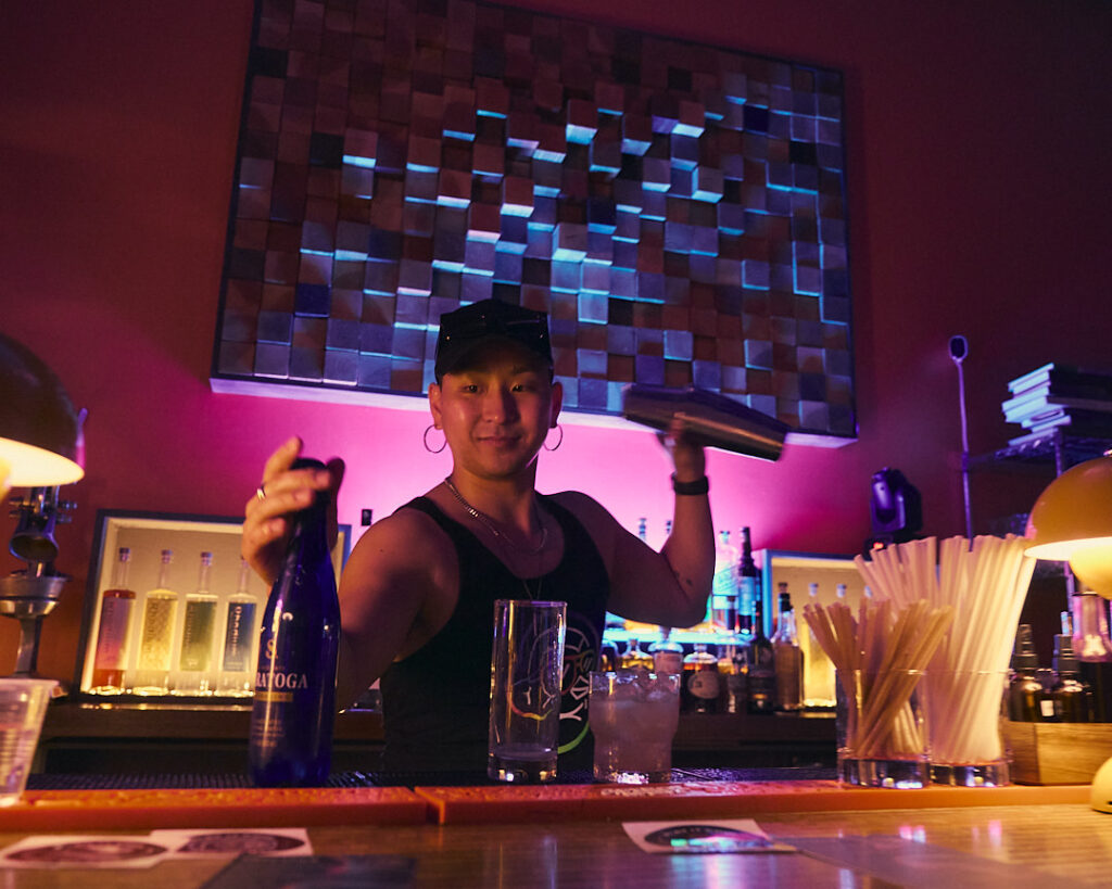 A bartender mixes a drink, holding a shaker and a bottle, in a colorful bar setting.