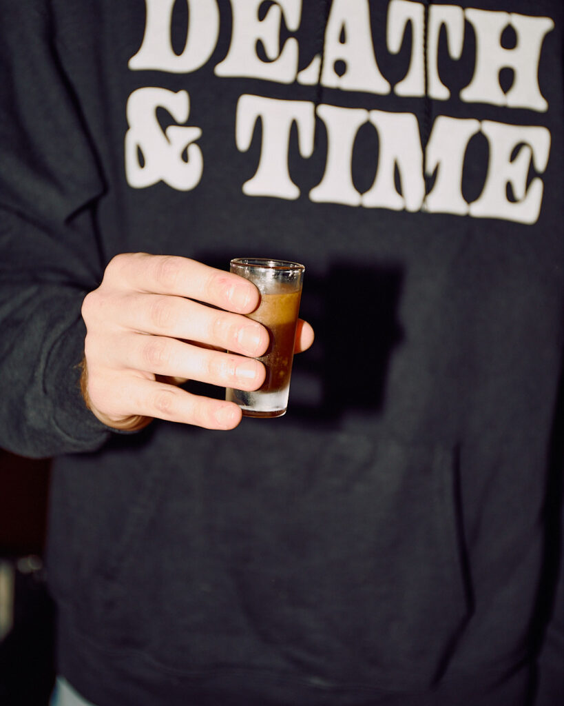A person holds a shot glass filled with a dark liquid, wearing a black hoodie with bold text.