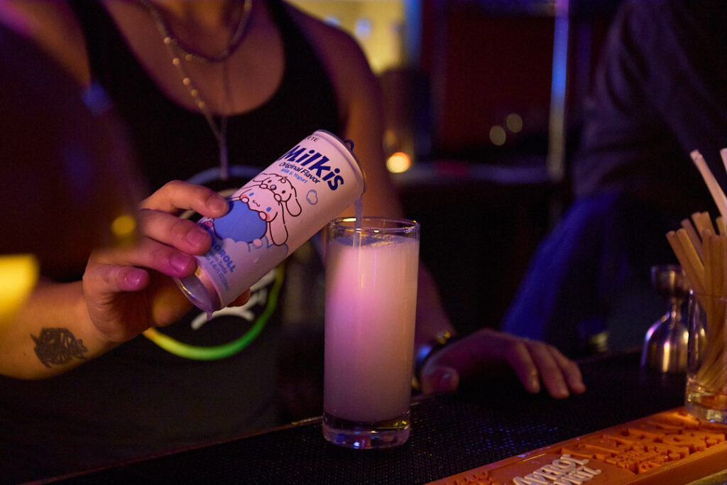 A person pours a drink from a can into a glass at a bar.