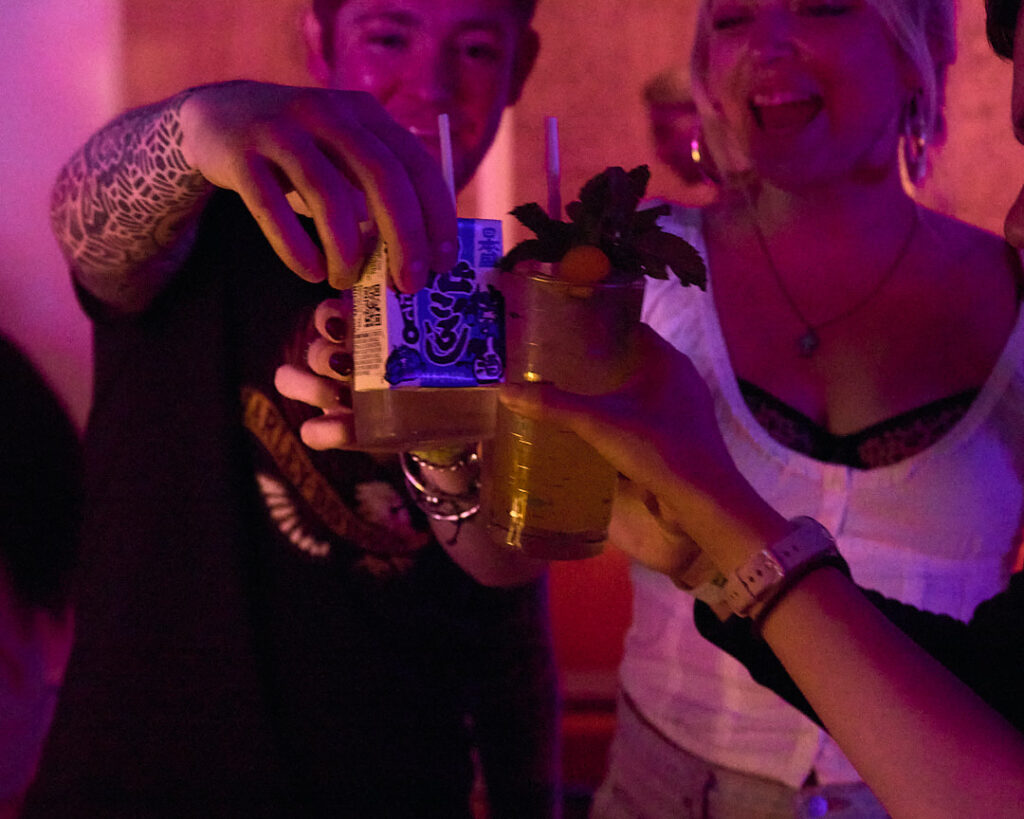 Friends toast with drinks, smiling and enjoying a lively atmosphere. Colorful lights illuminate the scene.