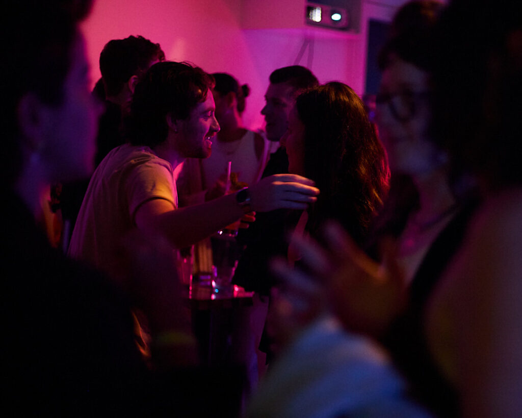 People socialize in a dimly lit space with colorful lighting, engaging in conversation and holding drinks.