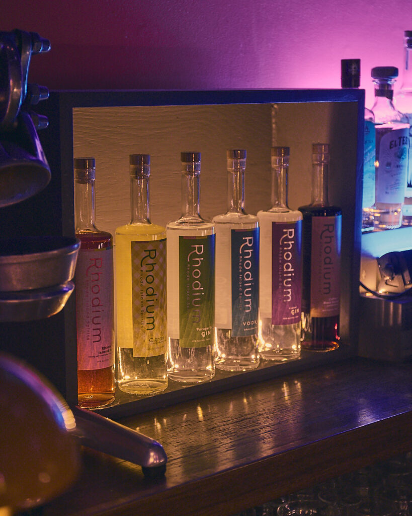 Five bottles of Rhodium spirits displayed on a shelf, with colorful labels under ambient lighting.