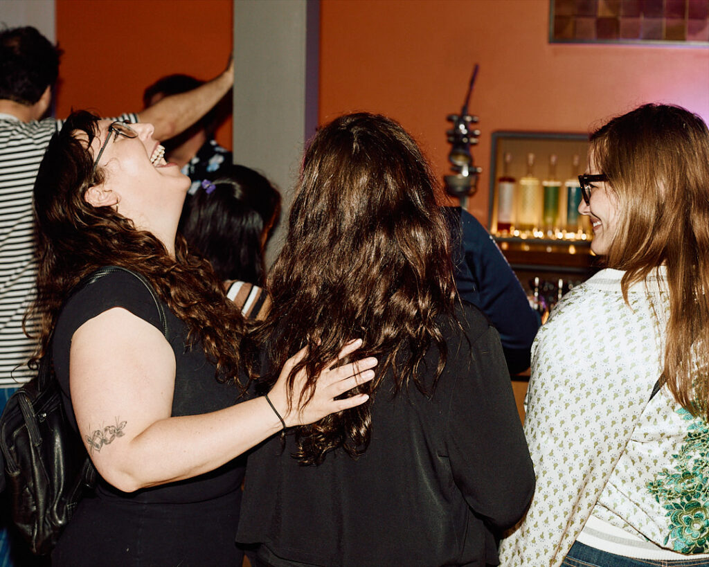 Three people laugh and interact in a lively bar setting, with colorful bottles in the background.
