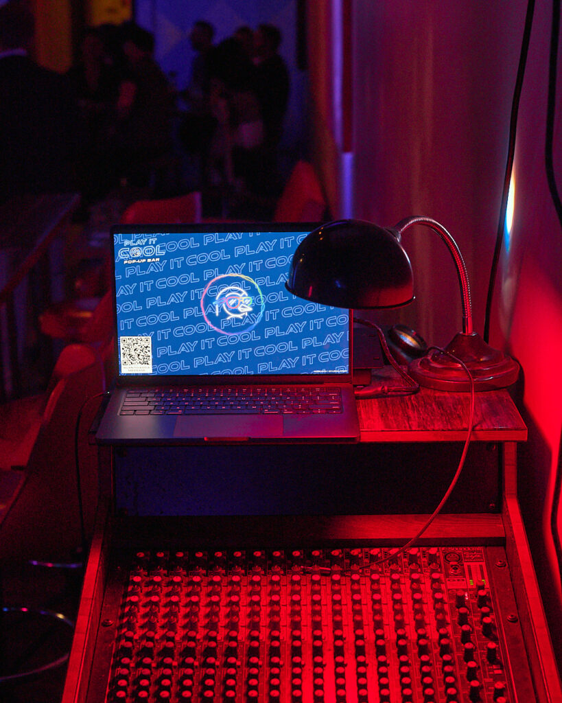 A laptop displays "PLAY IT COOL" on a dimly lit soundboard with a desk lamp. People are blurred in the background.