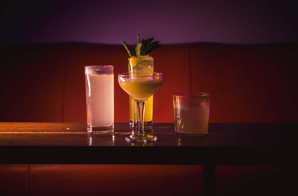Four craft cocktails displayed on a table at the "Craft Cocktail Play It Cool Pop-Up Bar Event" with a moody backdrop.