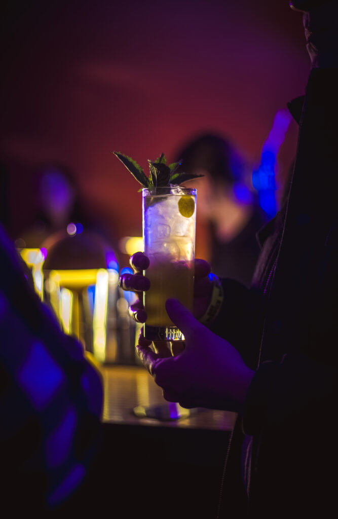 A person holds a cocktail with mint and lime at the Craft Cocktail Play It Cool Pop-Up Bar Event, with a dimly lit background.