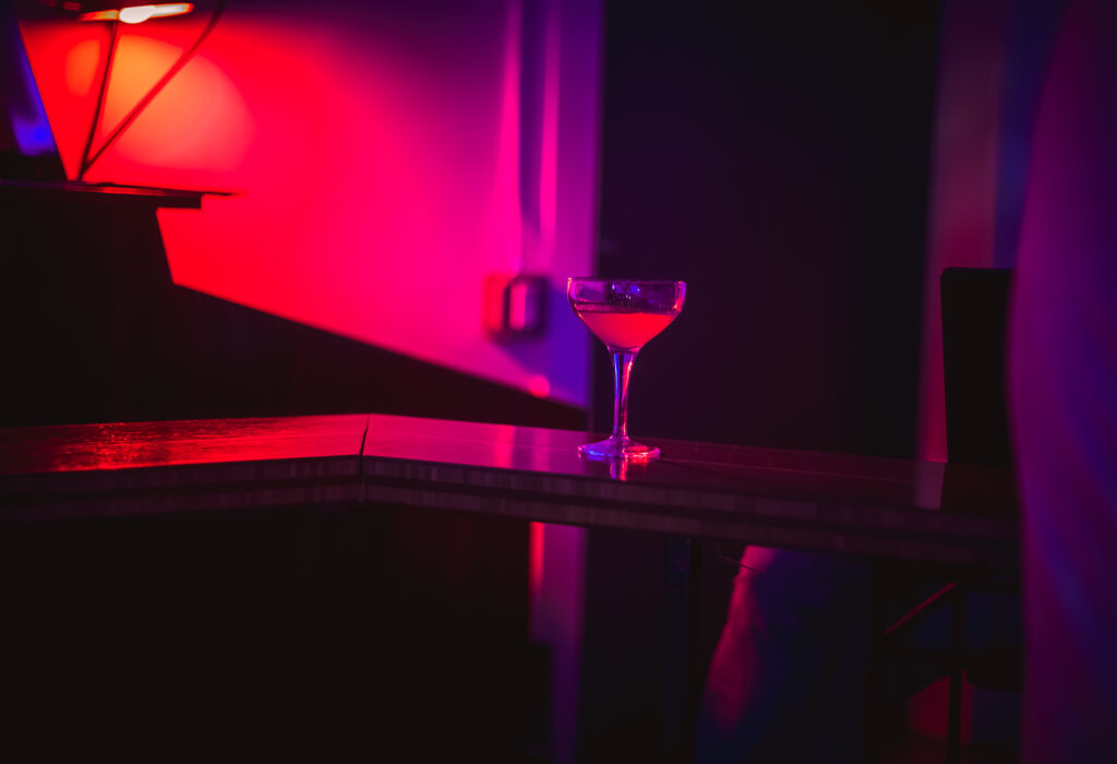 A cocktail glass on a bar counter at the Craft Cocktail Play It Cool Pop-Up Bar Event, illuminated by vibrant red and purple lighting.