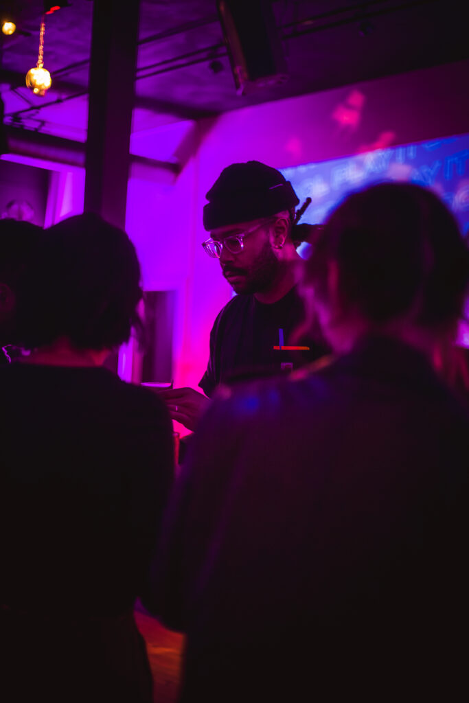 "Play It Cool Pop-Up Bar Event" features a person in a beanie and glasses in a dimly lit, purple-toned atmosphere.