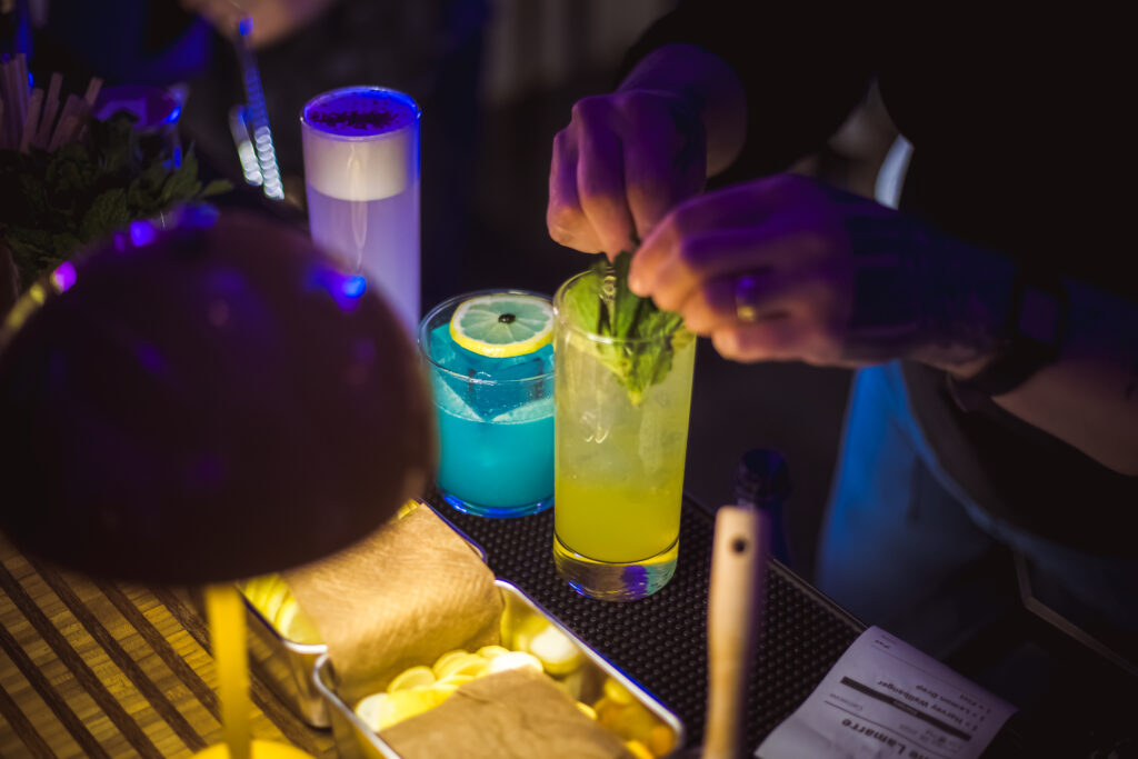A bartender garnishes a yellow cocktail with mint at the "Play It Cool Pop-Up Bar Event," surrounded by colorful drinks.