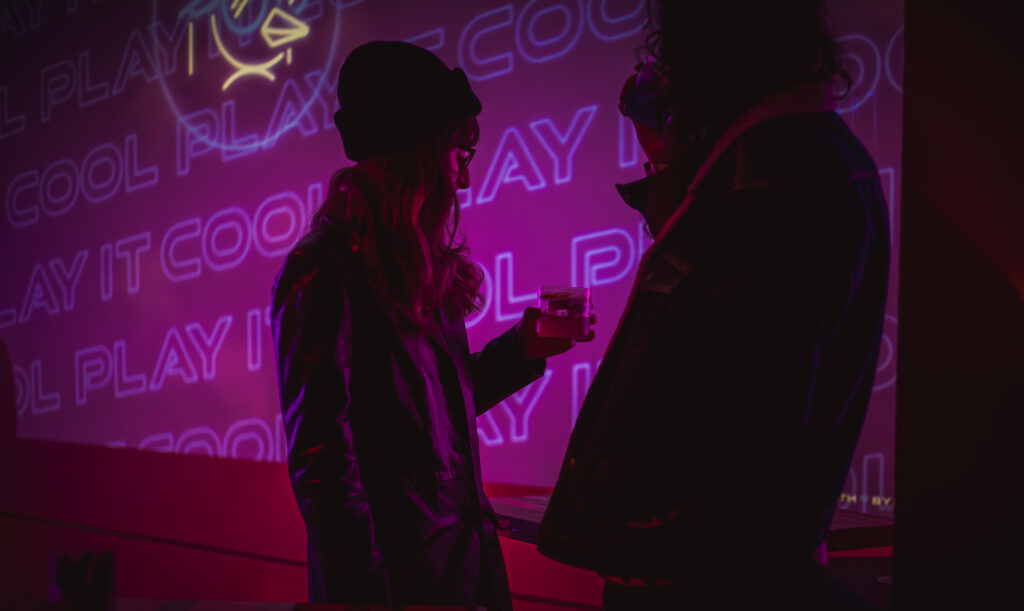 Two people at the "Play It Cool Pop-Up Bar Event" holding drinks, silhouetted against neon pink and purple lights.