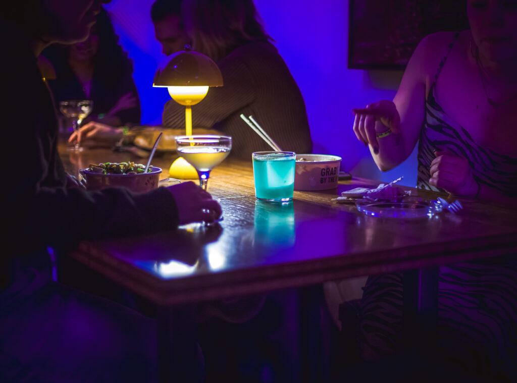 "Play It Cool Pop-Up Bar Event" features a dimly lit table with colorful drinks, snacks, and a stylish yellow lamp.