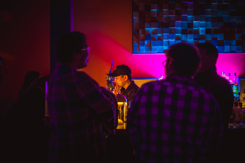 Play It Cool Founder Parker Luthman at a dimly lit bar, surrounded by people, with vibrant pink and blue lighting in the background.