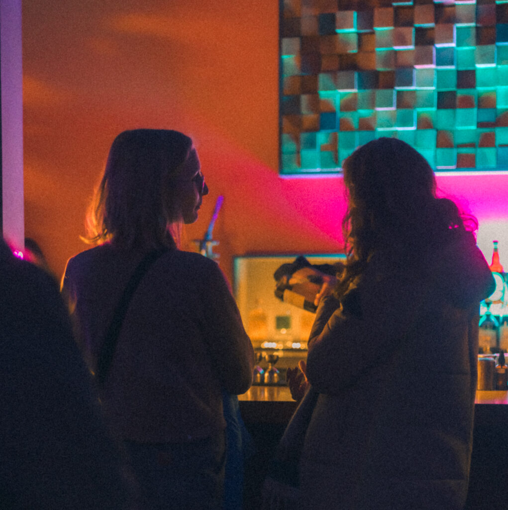 Two people conversing at the "Play It Cool Pop-Up Bar Event" with colorful lighting in the background.