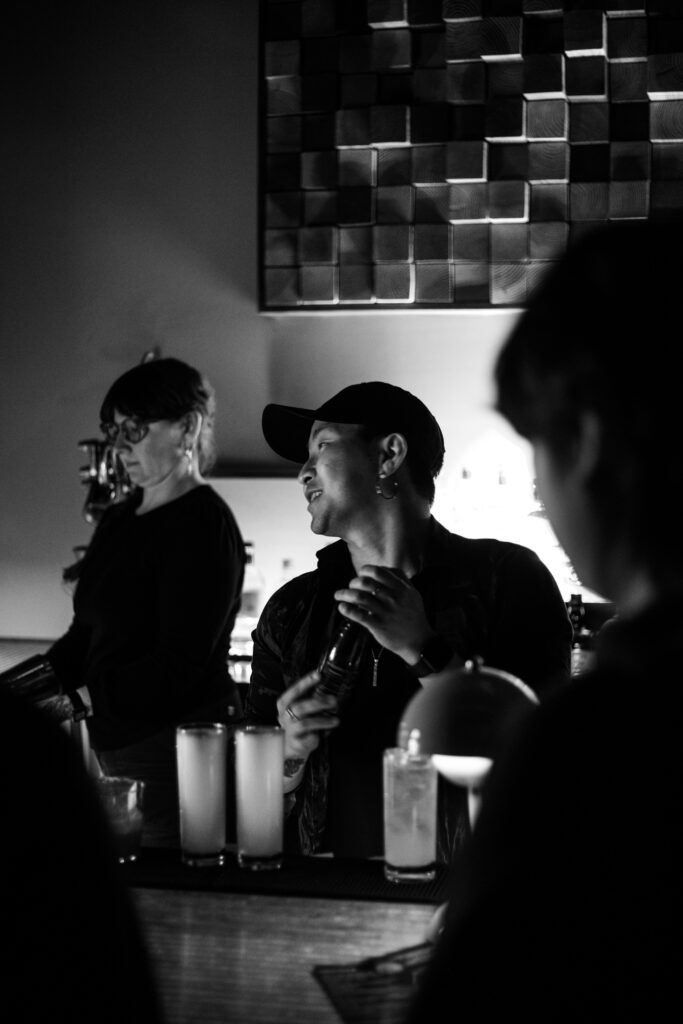 Play It Cool Founder Parker Luthman, wearing a cap, stands behind a bar, holding a shaker with drinks in the foreground.