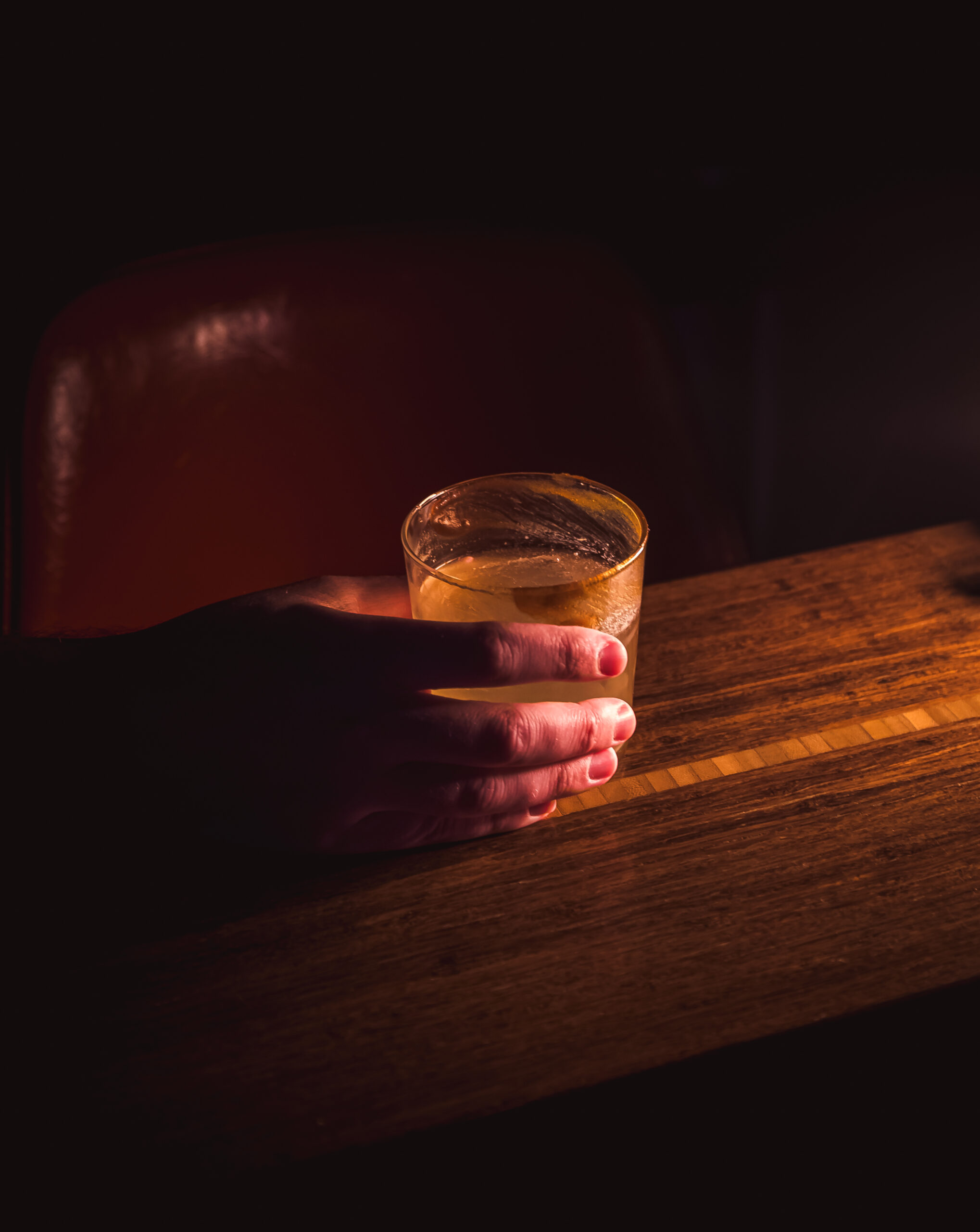 A hand holds a glass of Old Fashioned cocktail on a wooden table in low light.