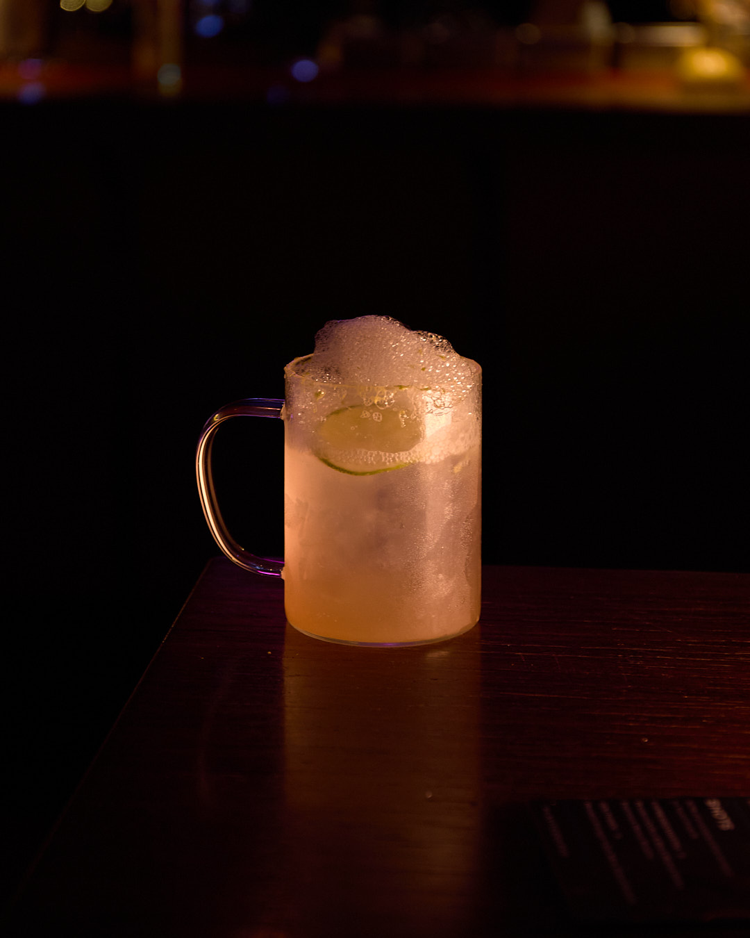 A frothy vodka soda in a glass mug with a lime slice, resting on a dark wooden table.