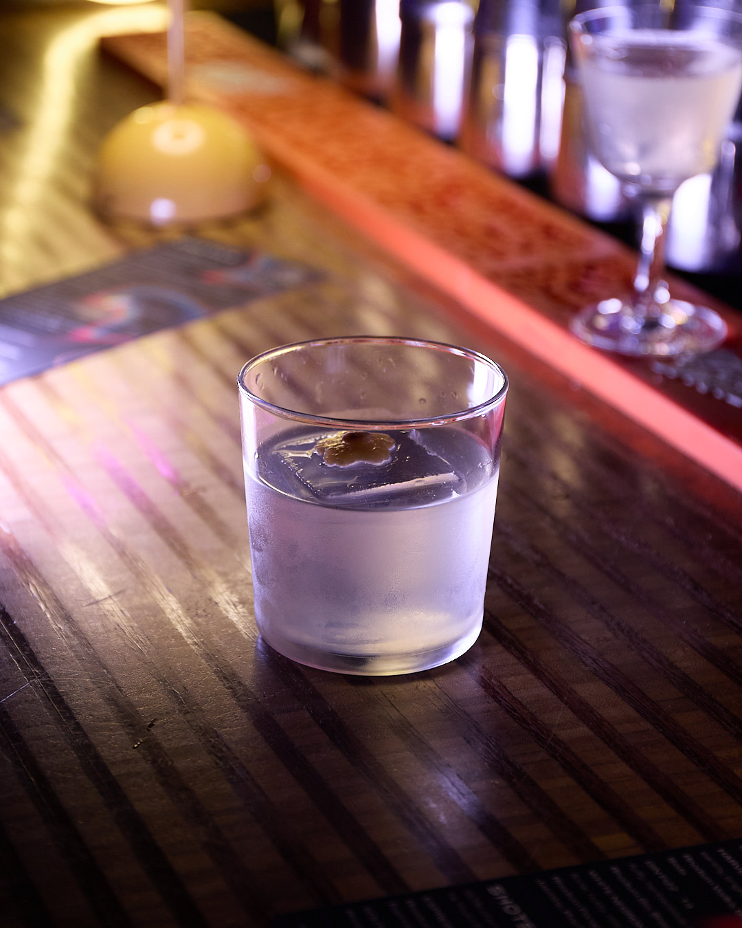 A glass of White Negroni with ice sits on a wooden bar, illuminated by soft lighting.